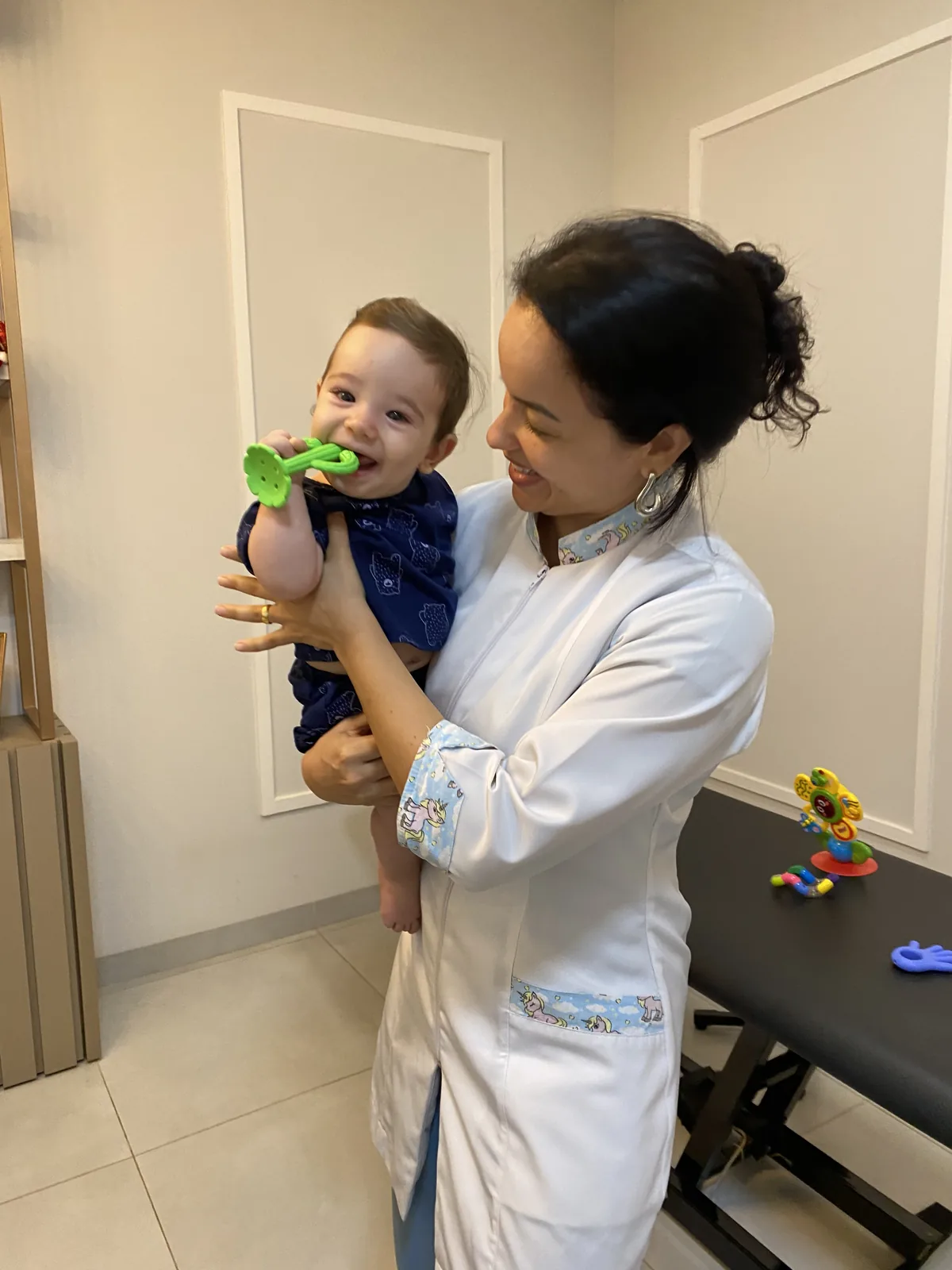 Dra. Ligiane Gama sorrindo durante atendimento de osteopatia pediátrica com um bebê no colo, em consultório em Sinop