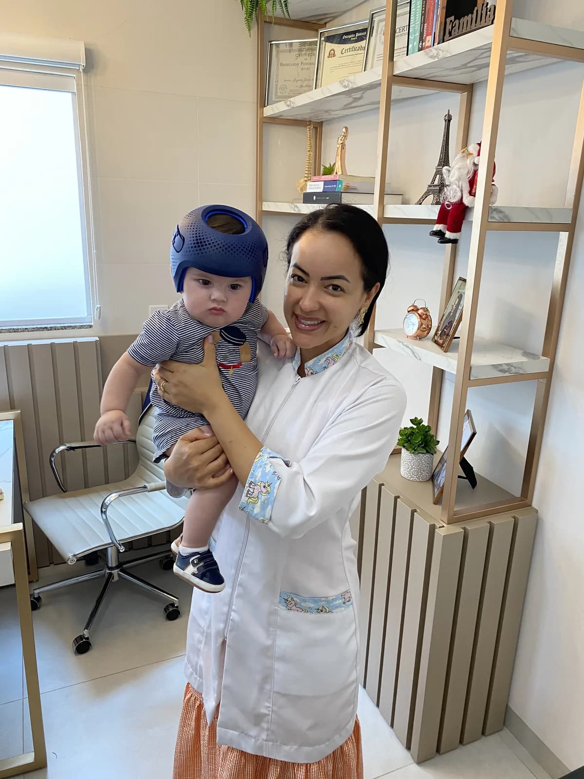 Dra. Ligiane Gama em seu consultório em Sinop, com um bebê usando capacete craniano no colo e certificados ao fundo